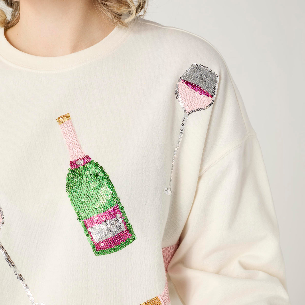 Cheers Sequin Sweatshirt, A smiling woman in an ivory sweatshirt covered in sequin champagne bottles and wine glasses in pink, green and silver, with blue jeans.