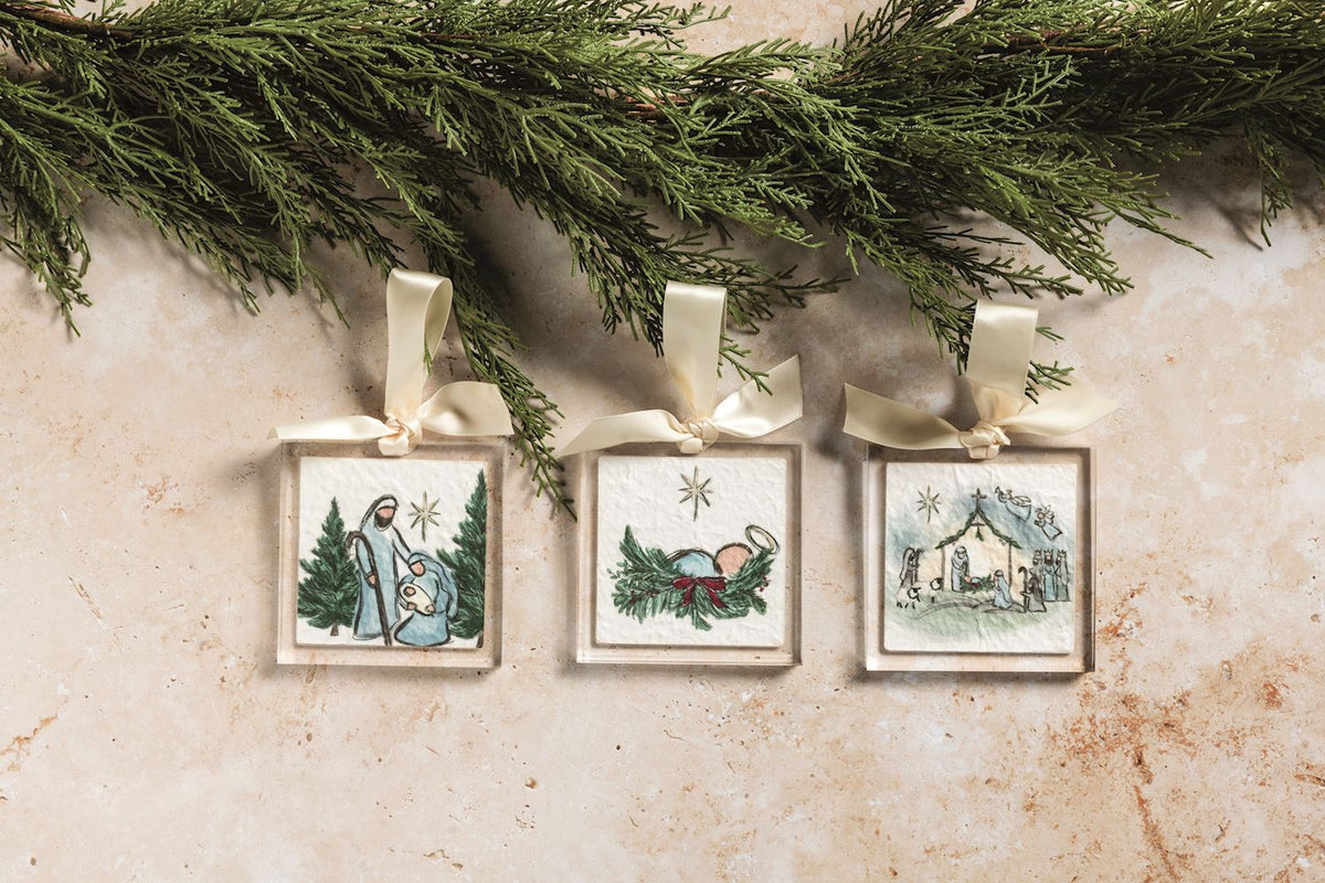 Mary Nativity Acrylic Ornament, Square clear acrylic ornament holding a hand drawn Nativity scene of the Holy Family under a star between two evergreen trees, hung from a cream grosgrain ribbon bow.