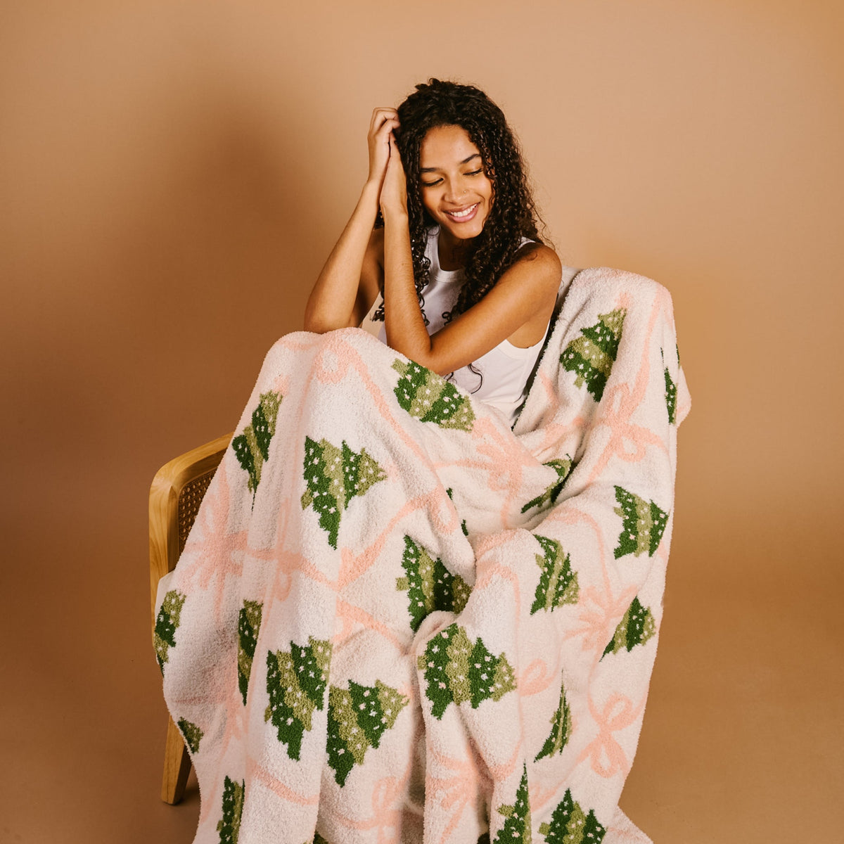 Twinkling Trees Cozy Dreams Luxe Blanket, A cream throw blanket printed with a pattern of green Christmas trees connected by a pink ribbon lattice, held up to show the full design.