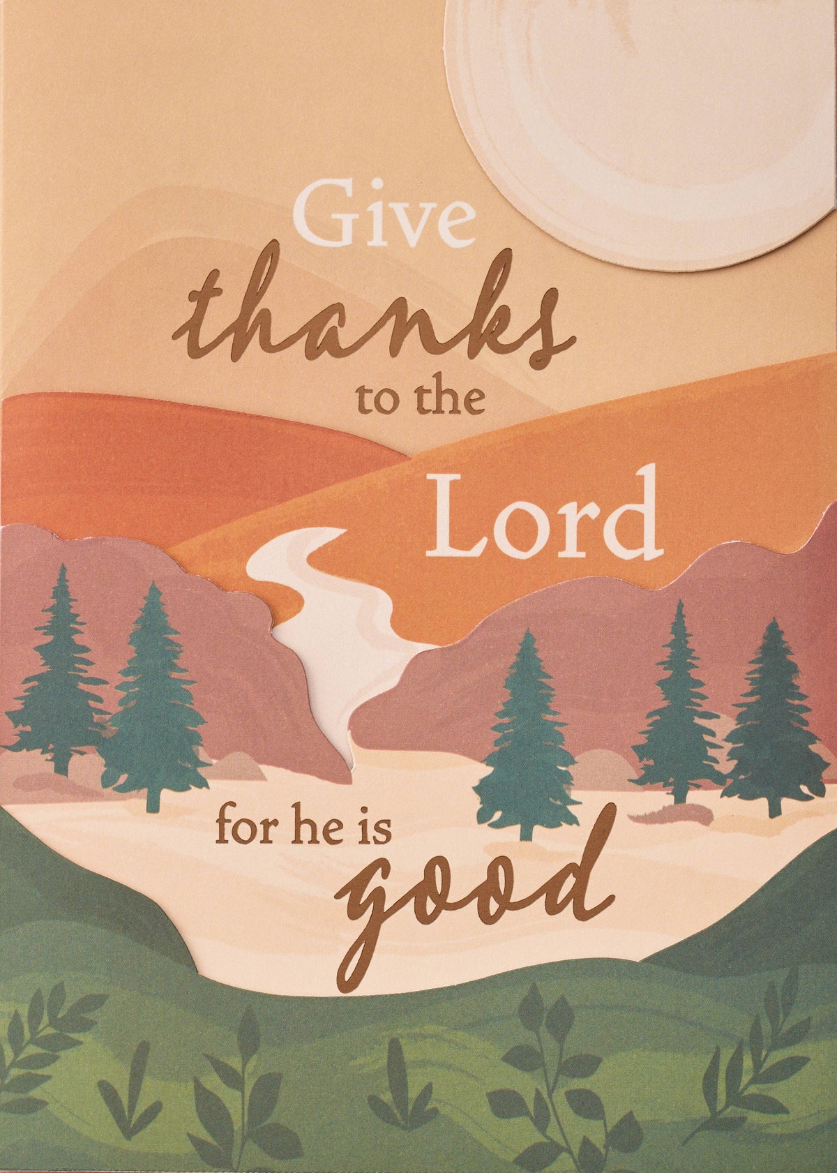 Divine Gratitude Greeting Card, A card front with a landscape of stacked peach, orange and rose hills, evergreen trees, a winding pale river and a round cream sun, with the words Give thanks to the Lord for he is good in brown script and cream lettering.