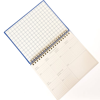 Cobalt Blue Fabric-Bound Weekly Desk Pad, A cobalt blue fabric desk pad reading days weeks in gold foil with a spiral top binding.