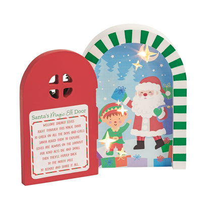 Light-Up Wooden Magic Elf Door, 5 Inch, An arched wooden elf door with a red front panel, a cut-out window and a printed verse, standing in front of a painted winter scene of Santa and an elf lit by small stars.
