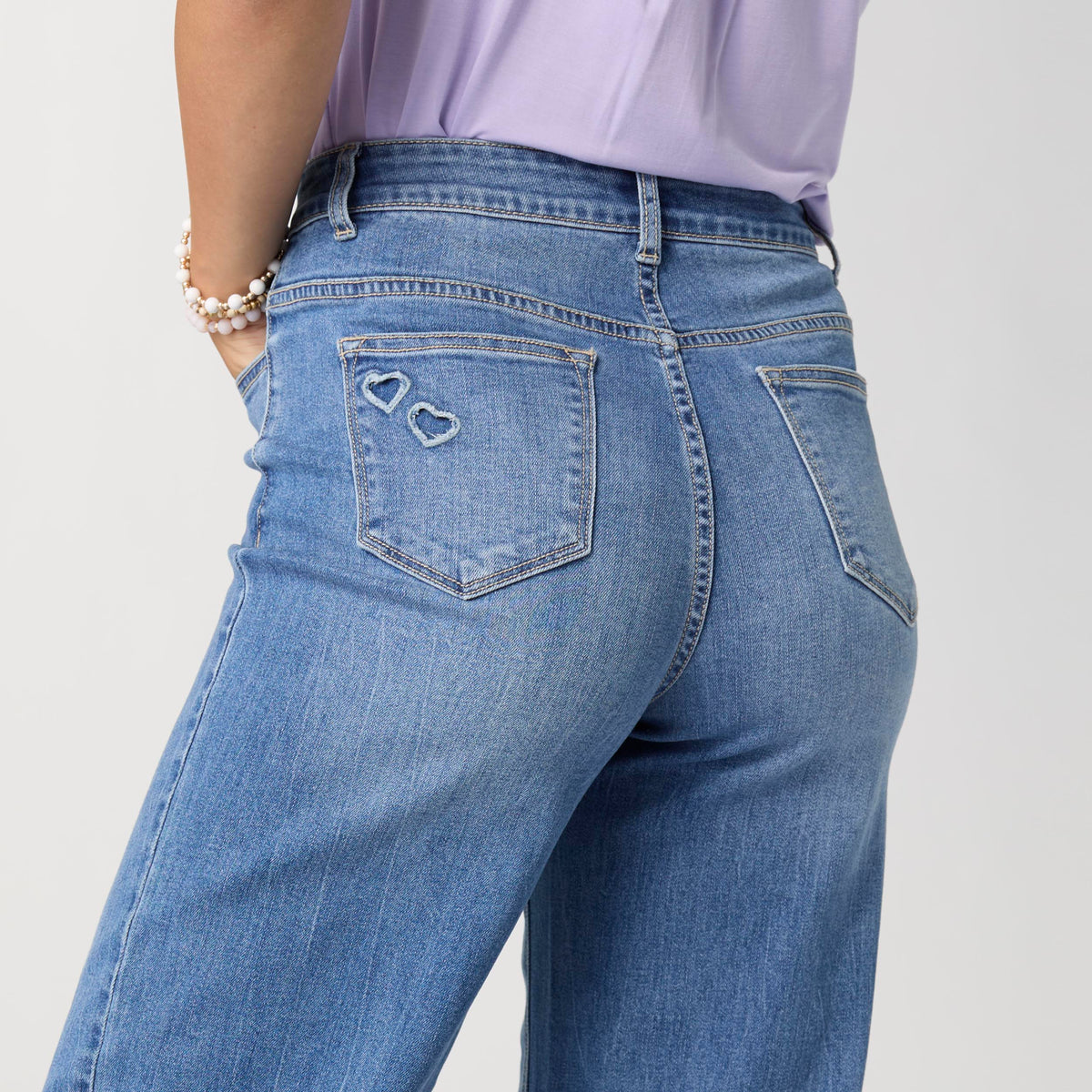 Bambu Soft Heart Cut-Out Wide Leg Ankle Jeans, A woman in medium denim wide leg ankle jeans with heart cut-outs along the hems, a lavender top and braided sandals.