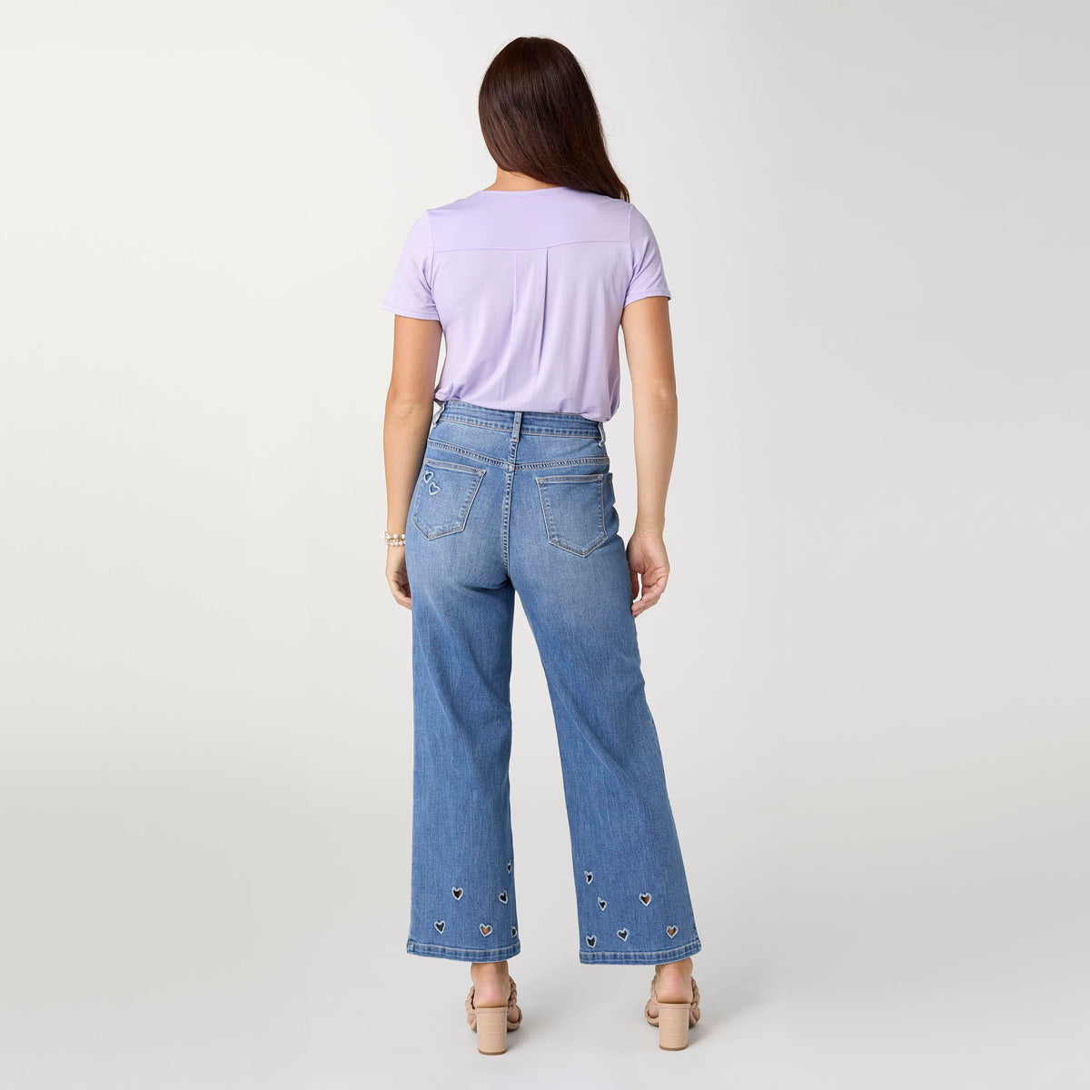 Bambu Soft Heart Cut-Out Wide Leg Ankle Jeans, A woman in medium denim wide leg ankle jeans with heart cut-outs along the hems, a lavender top and braided sandals.