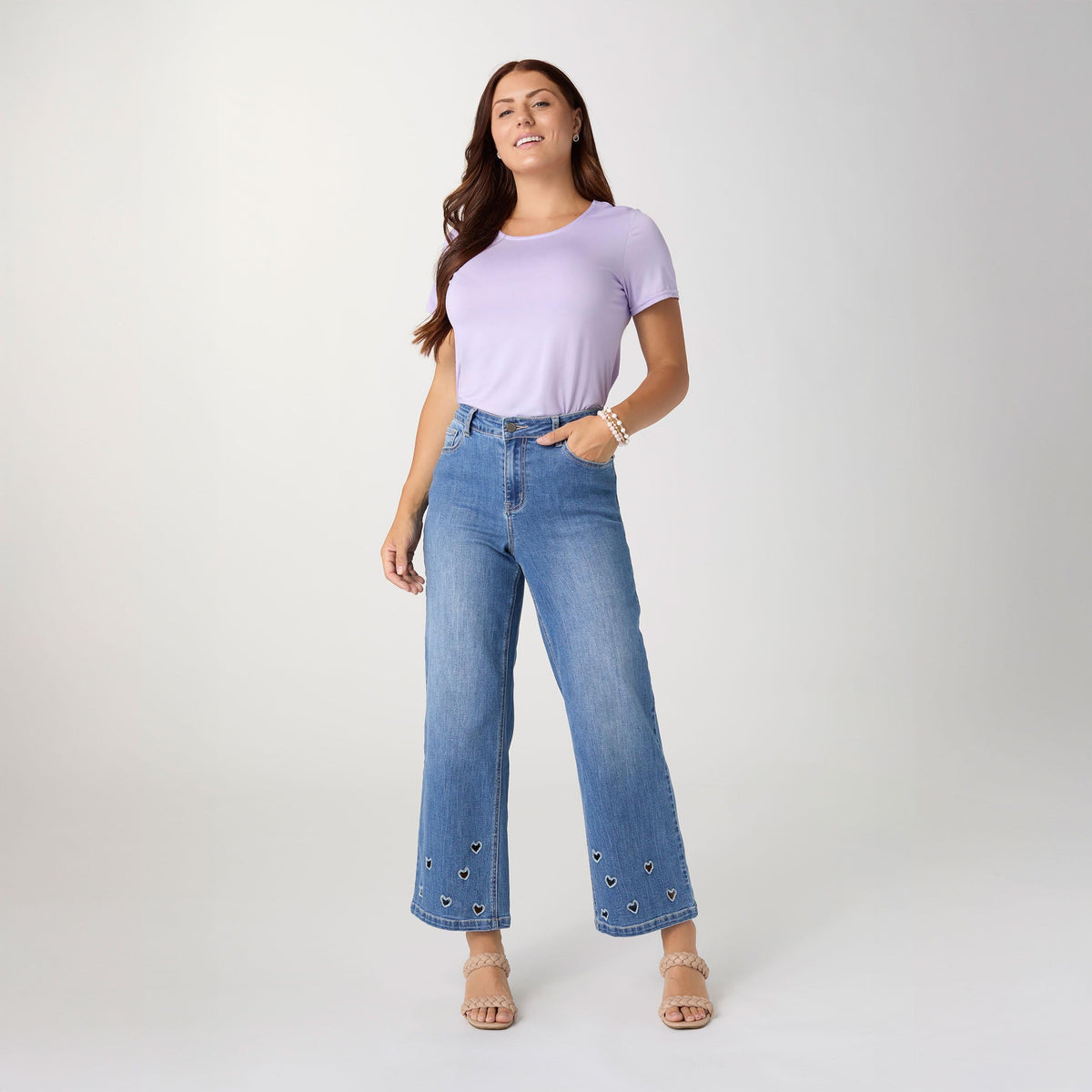 Bambu Soft Heart Cut-Out Wide Leg Ankle Jeans, A woman in medium denim wide leg ankle jeans with heart cut-outs along the hems, a lavender top and braided sandals.