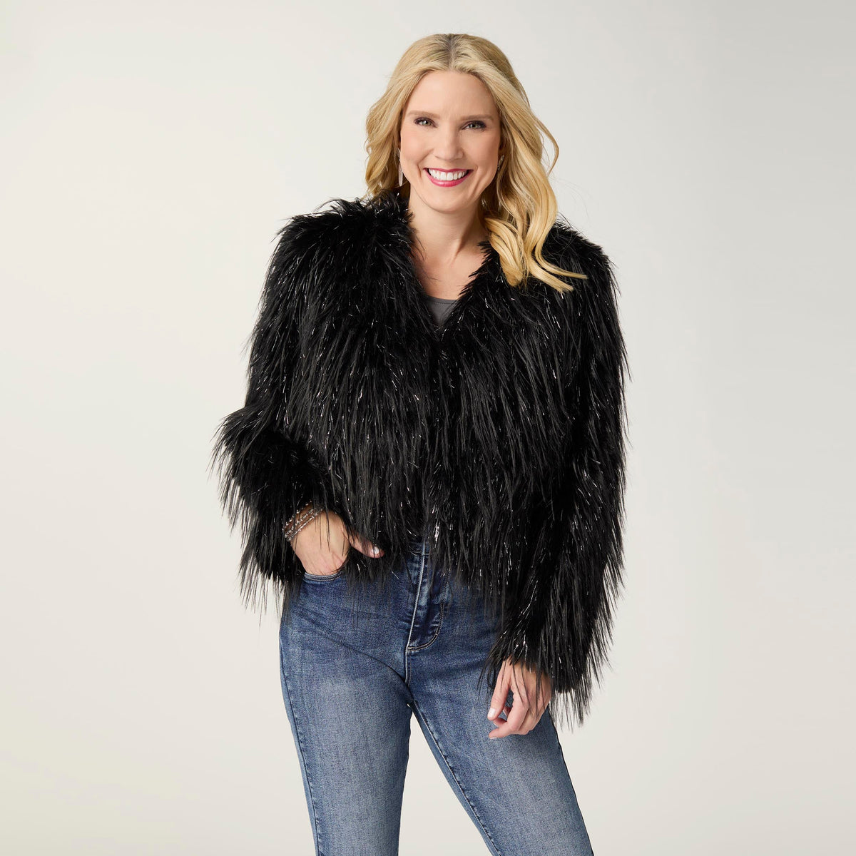 Raven Tinsel Faux Fur Jacket, A smiling woman in a cropped black faux fur jacket with silver tinsel strands over a gray top and blue jeans.