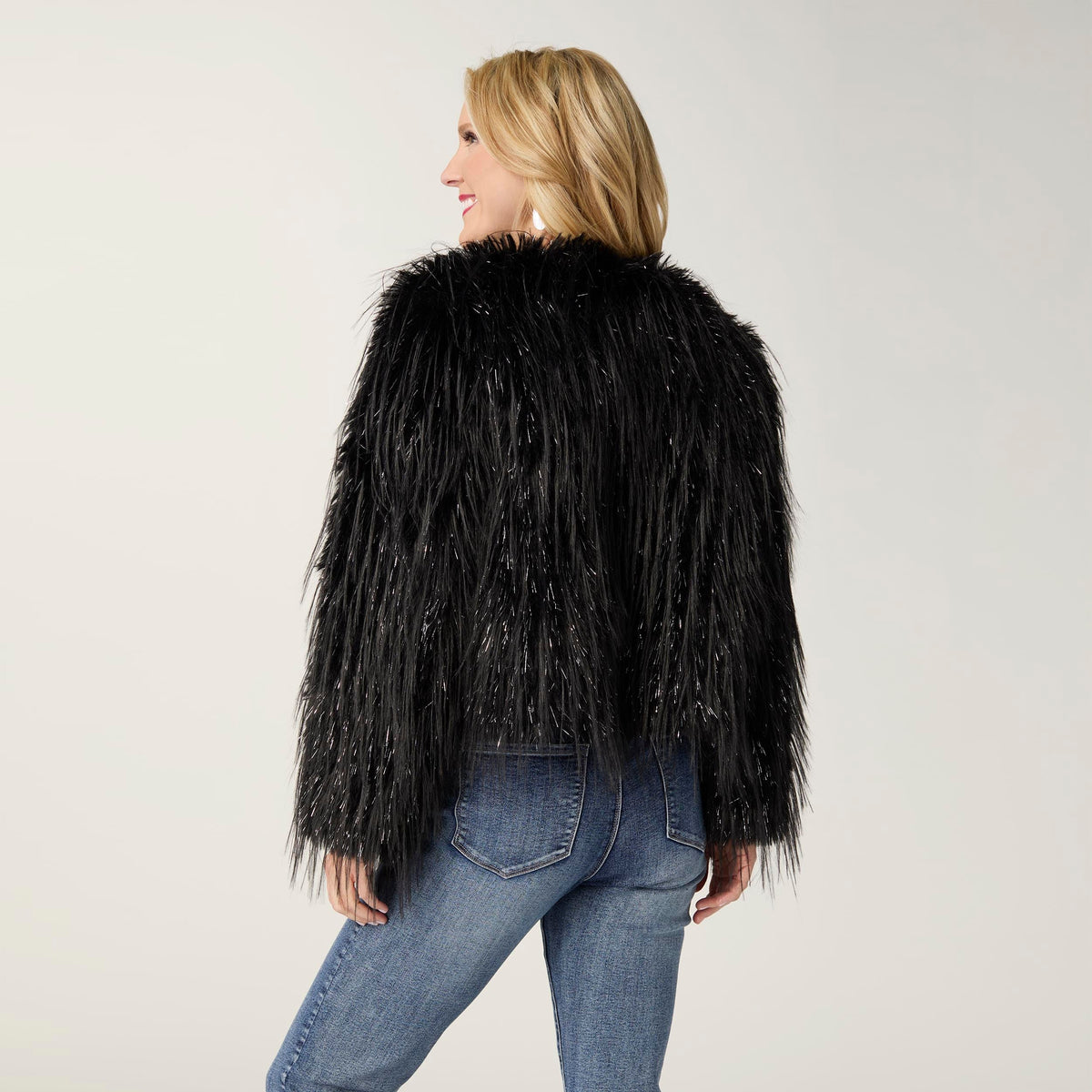 Raven Tinsel Faux Fur Jacket, A smiling woman in a cropped black faux fur jacket with silver tinsel strands over a gray top and blue jeans.
