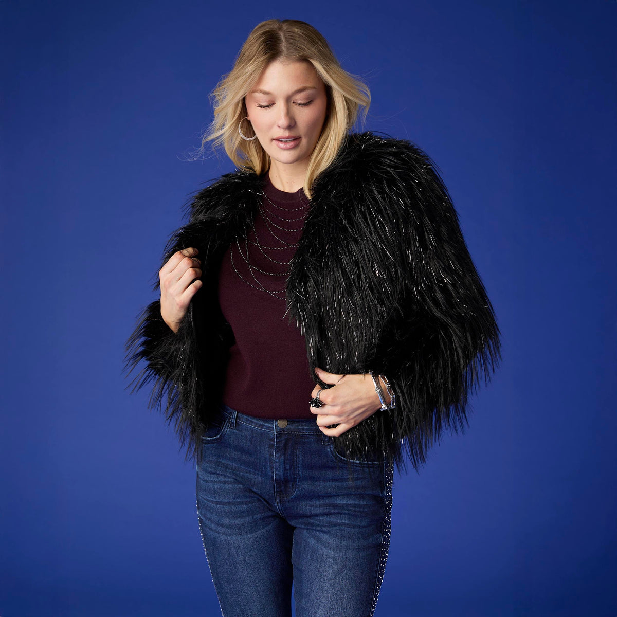 Raven Tinsel Faux Fur Jacket, A smiling woman in a cropped black faux fur jacket with silver tinsel strands over a gray top and blue jeans.