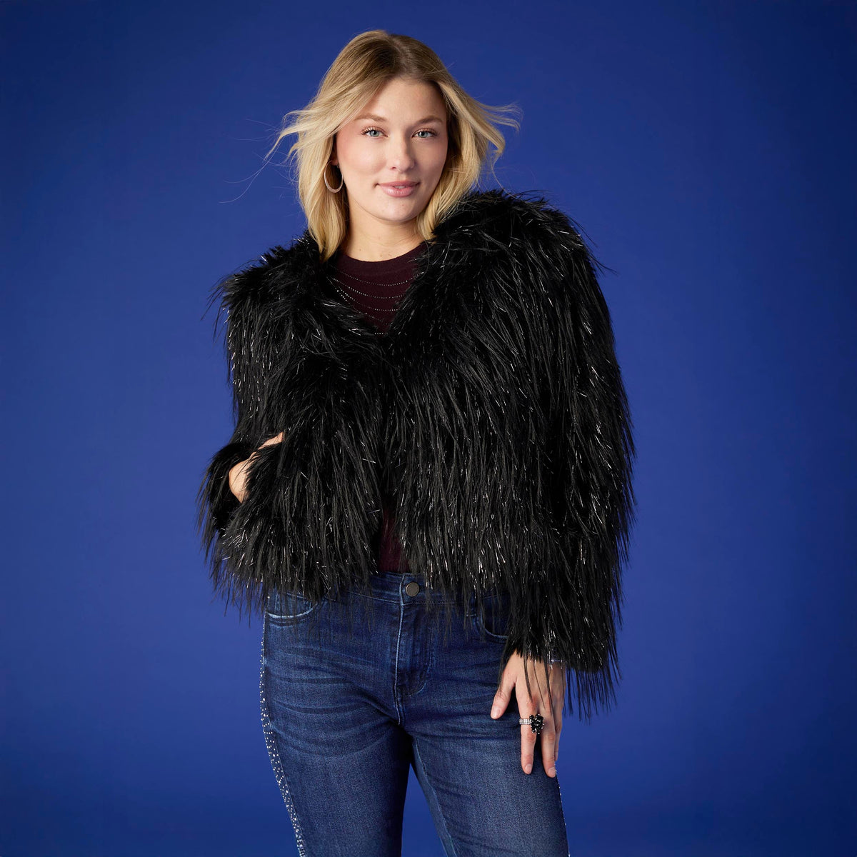 Raven Tinsel Faux Fur Jacket, A smiling woman in a cropped black faux fur jacket with silver tinsel strands over a gray top and blue jeans.