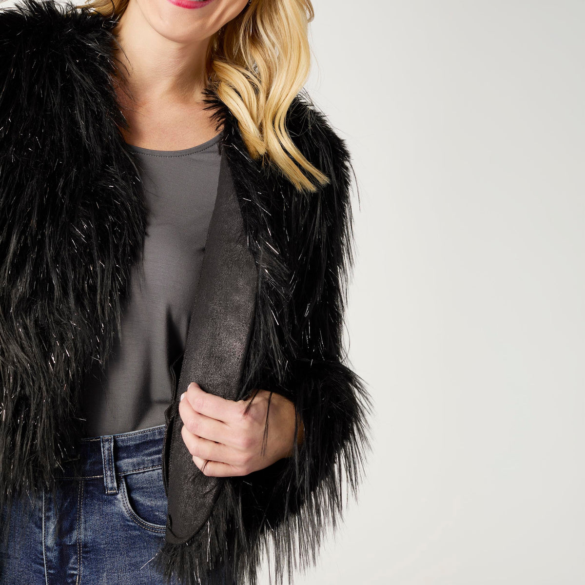 Raven Tinsel Faux Fur Jacket, A smiling woman in a cropped black faux fur jacket with silver tinsel strands over a gray top and blue jeans.