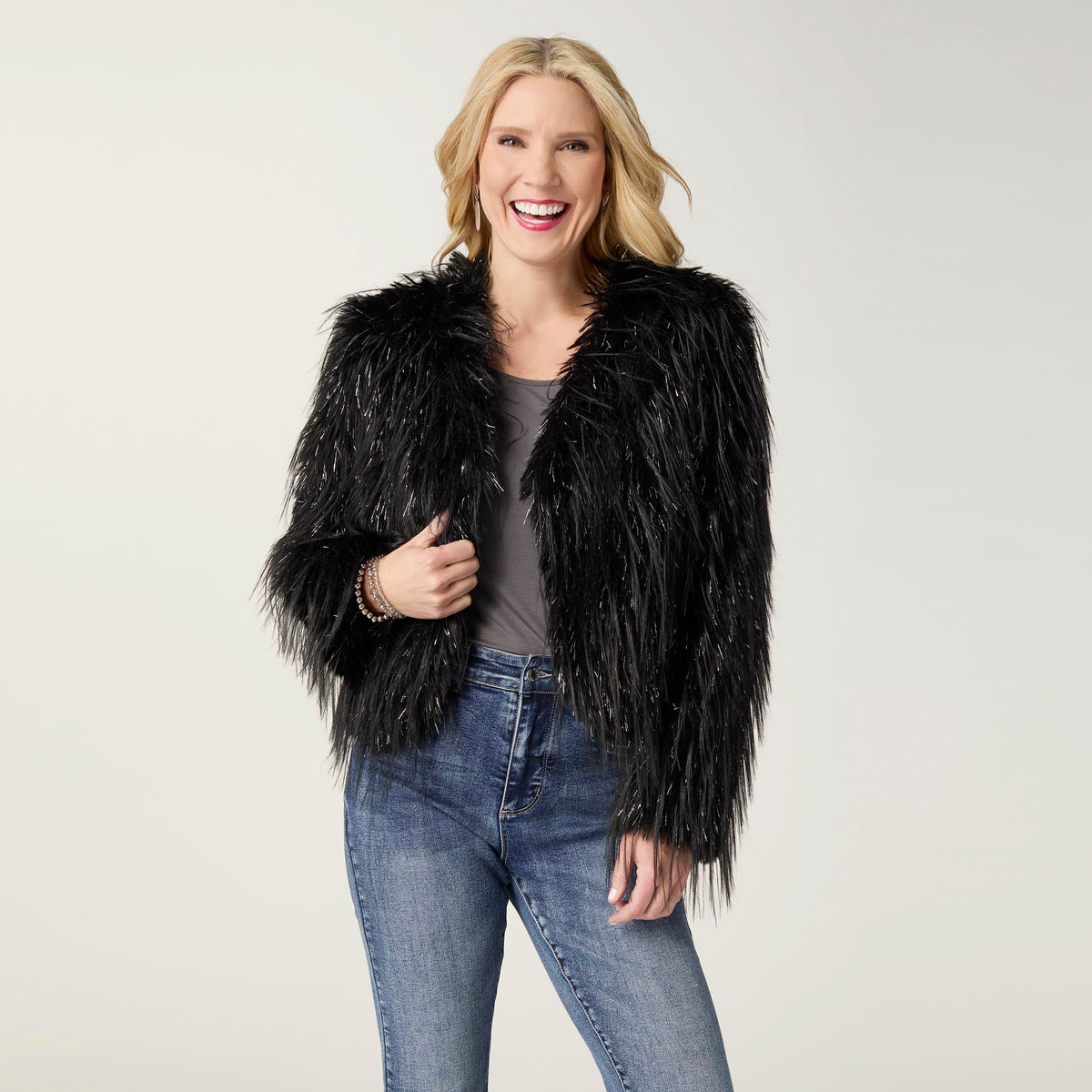 Raven Tinsel Faux Fur Jacket, A smiling woman in a cropped black faux fur jacket with silver tinsel strands over a gray top and blue jeans.