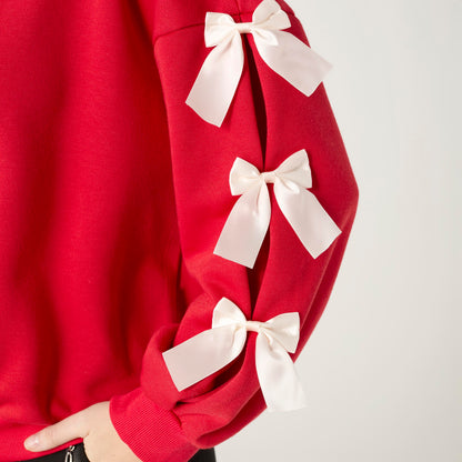 Pleated Bow Sleeve Sweatshirt, A smiling woman in a red crew neck sweatshirt with cream bows down one sleeve and black pants.