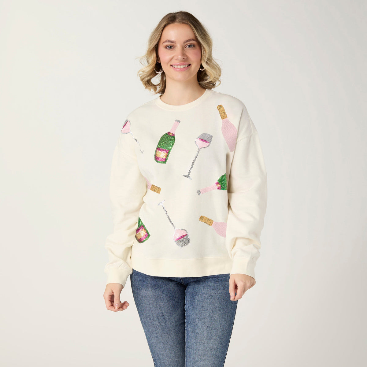 Cheers Sequin Sweatshirt, A smiling woman in an ivory sweatshirt covered in sequin champagne bottles and wine glasses in pink, green and silver, with blue jeans.