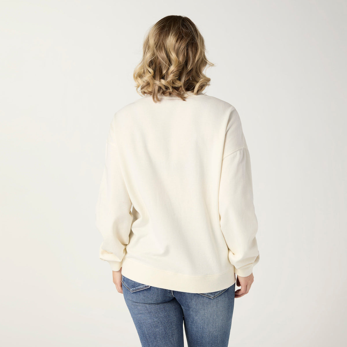 Cheers Sequin Sweatshirt, A smiling woman in an ivory sweatshirt covered in sequin champagne bottles and wine glasses in pink, green and silver, with blue jeans.