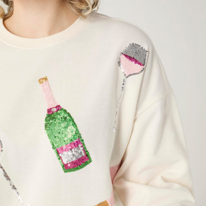 Cheers Sequin Sweatshirt, A smiling woman in an ivory sweatshirt covered in sequin champagne bottles and wine glasses in pink, green and silver, with blue jeans.
