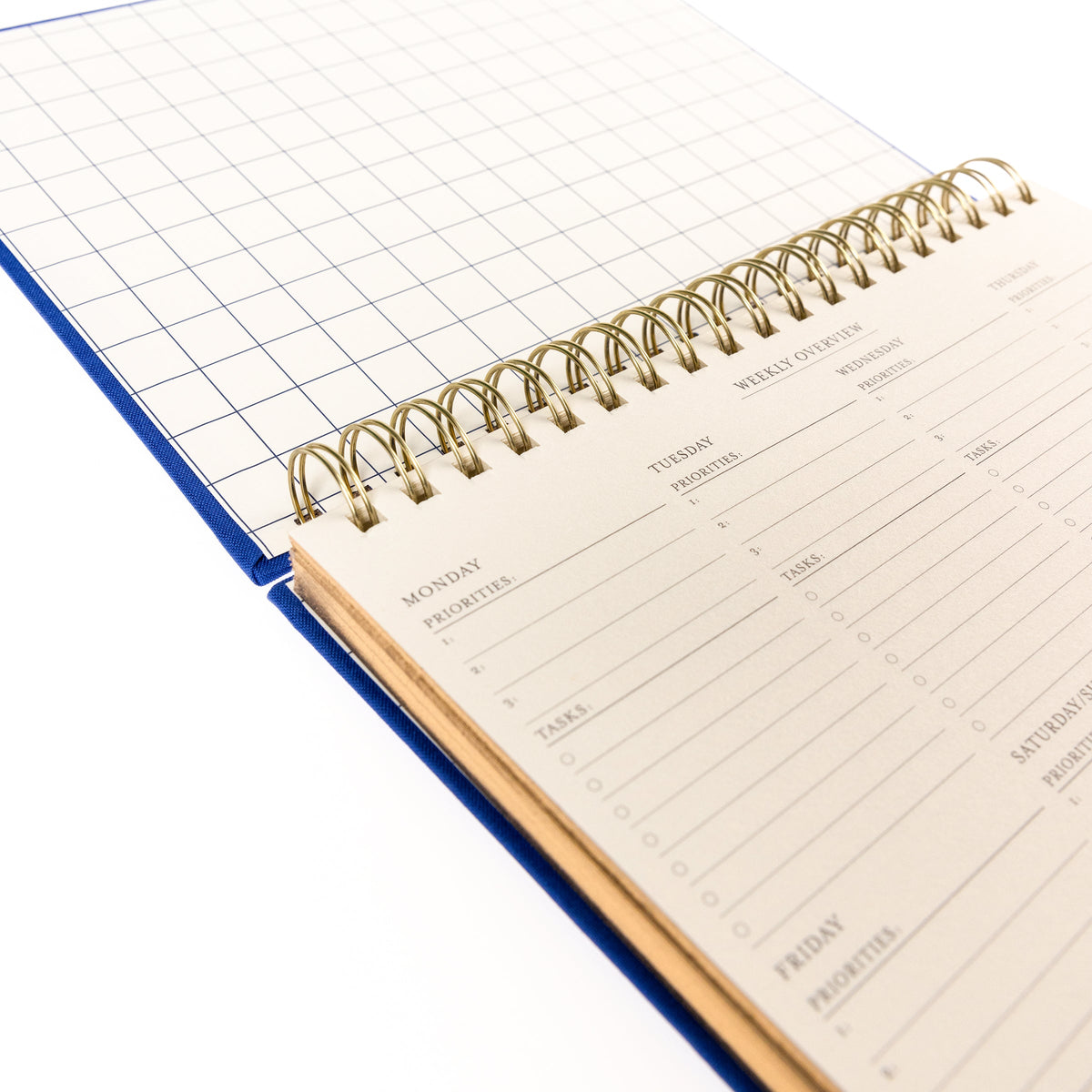 Cobalt Blue Fabric-Bound Weekly Desk Pad, A cobalt blue fabric desk pad reading days weeks in gold foil with a spiral top binding.