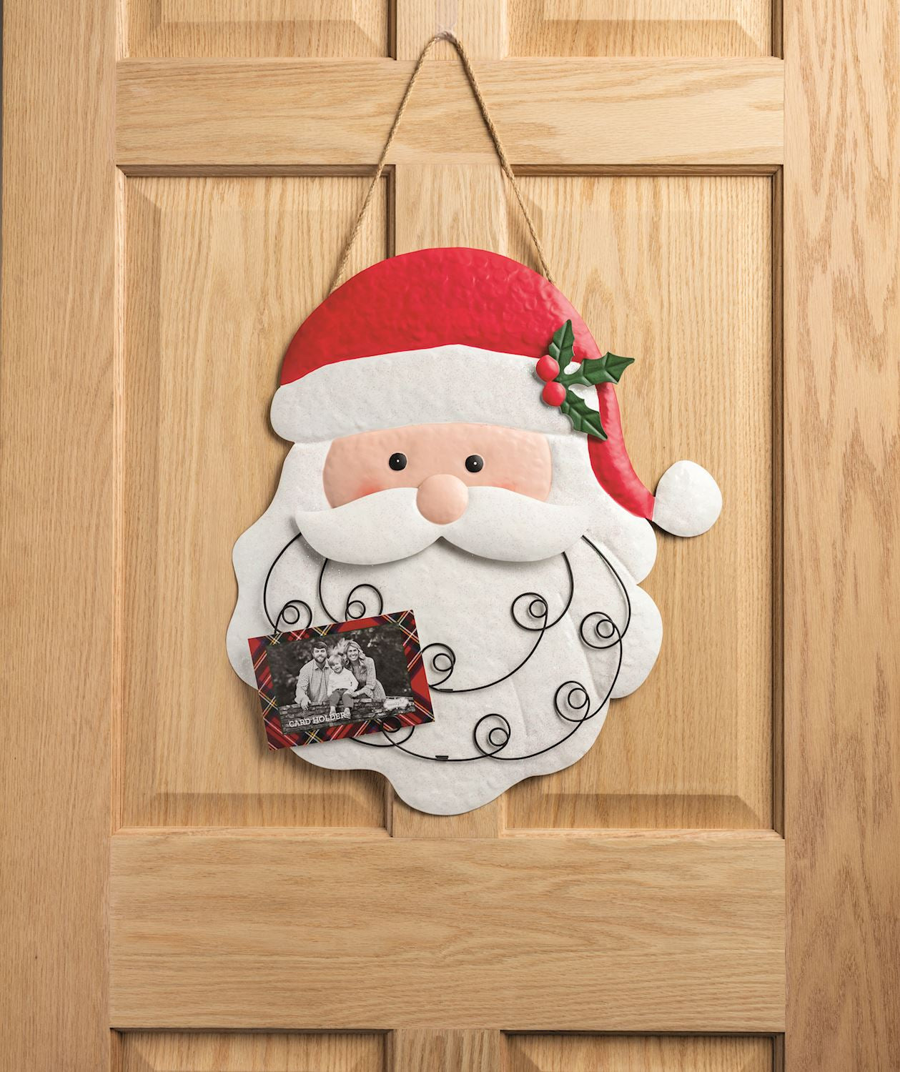Tin Santa Christmas Card Holder Wall Hanging, 20 Inch, A large tin Santa face with a red hat and holly sprig, a white beard crossed by black wire coils for holding cards, hanging from a jute rope.