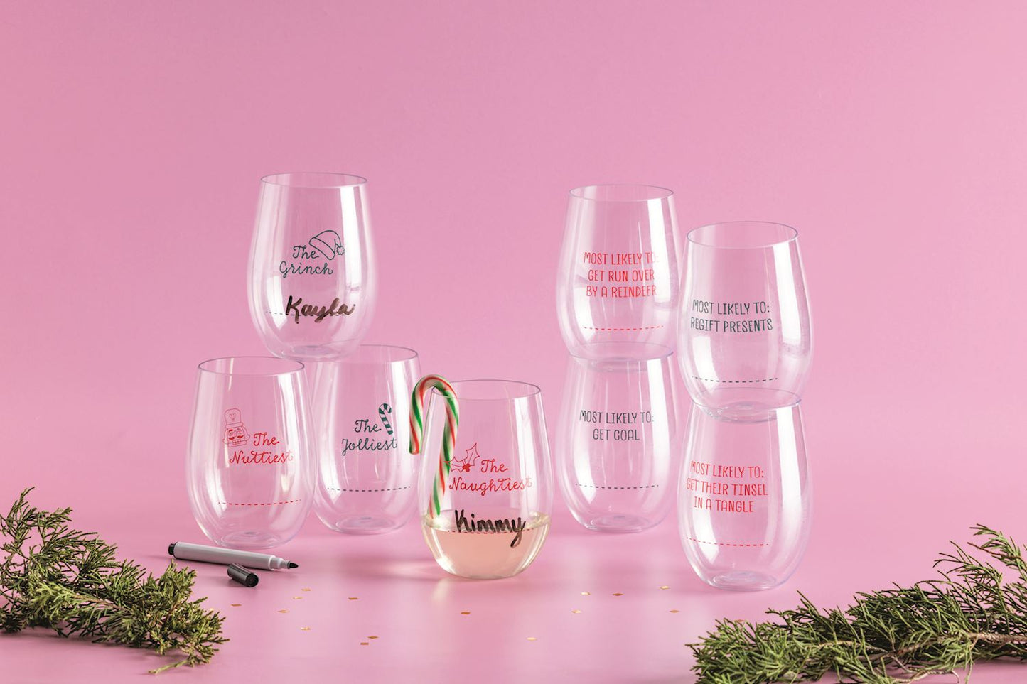Christmas Stemless Wine Cup Set with Marker, Four clear stemless wine cups stacked inside a cello sleeve tied with a green ribbon, each printed with a red or green holiday title and a dotted name line, with a black marker alongside.