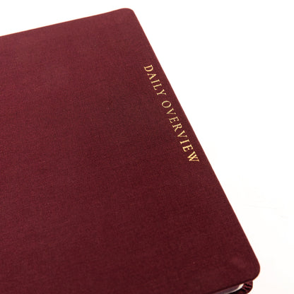Burgundy Fabric-Bound Task Journal, A burgundy fabric-covered task journal with gold twin wire spirals and two maroon ribbon bookmark tabs, closed with an elastic band.