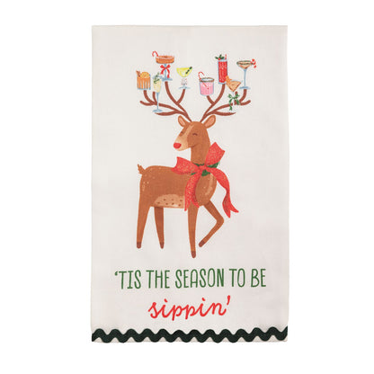 Tis the Season Reindeer Glitter Hand Towel, A cream cotton hand towel printed with a brown reindeer in a glittered red bow carrying holiday cocktails on his antlers, over the words tis the season to be sippin, with a dark green ric-rac hem.