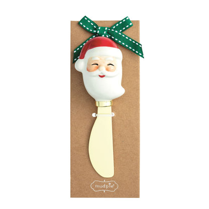 Santa Handle Cheese Spreader, 5.5 Inch, A brass-toned spreader with a hand-painted Santa head handle in a red cap with a white beard, mounted on a kraft card with a green ribbon with white stitching.