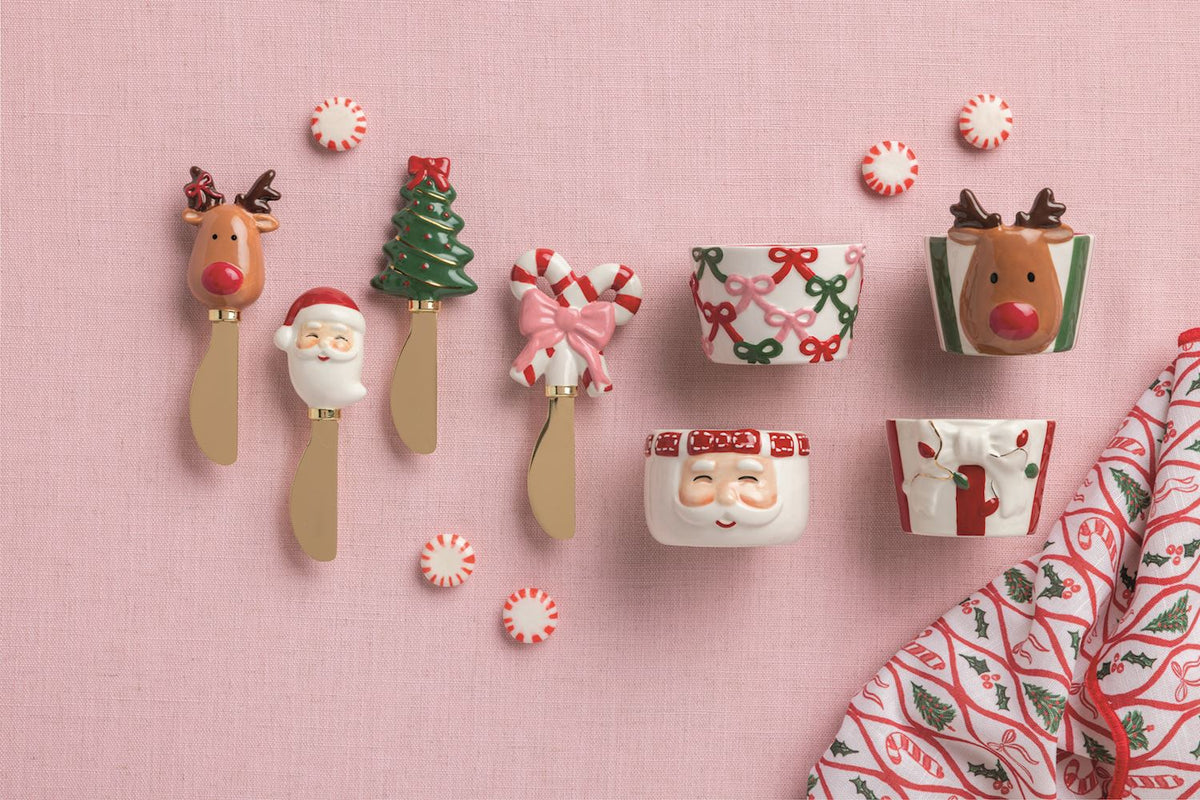 Santa Handle Cheese Spreader, 5.5 Inch, A brass-toned spreader with a hand-painted Santa head handle in a red cap with a white beard, mounted on a kraft card with a green ribbon with white stitching.