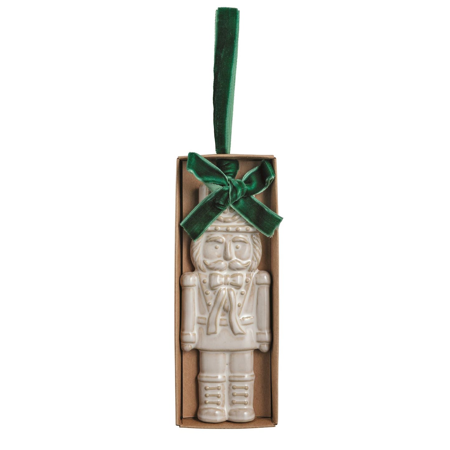 White Nutcracker Stoneware Figural Ornament, Cream glazed stoneware nutcracker ornament with its uniform pressed into the clay, tied with a green velvet ribbon bow and loop, lying in a small kraft gift box.