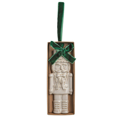 White Nutcracker Stoneware Figural Ornament, Cream glazed stoneware nutcracker ornament with its uniform pressed into the clay, tied with a green velvet ribbon bow and loop, lying in a small kraft gift box.