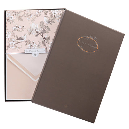 La Petite Presse Birds Boxed Correspondence Notes, A brown gift box open to a blush pink notecard printed with songbirds perched among magnolia blossoms, paired with a matching blush envelope.