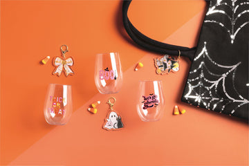 Lets Go Ghouls Wine Cup and Bag Charm Set, A clear stemless plastic wine cup printed with a haunted house, ghost, bats and pumpkin, with a pink and black lets go ghouls acrylic bag charm hanging inside it on a gold clip.