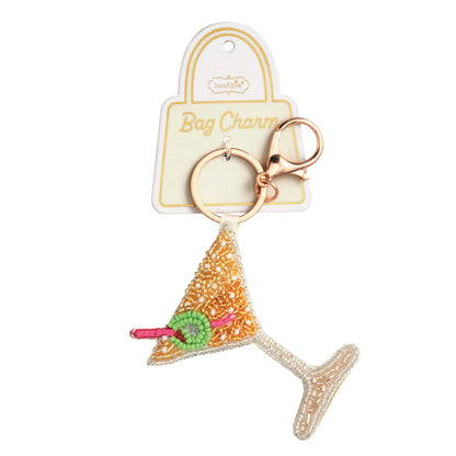Martini Beaded Bag Charm, A hand-beaded martini glass charm in gold beads with a clear beaded stem and a green olive on a pink pick, hanging from a gold key ring and lobster clasp on a Bag Charm card.
