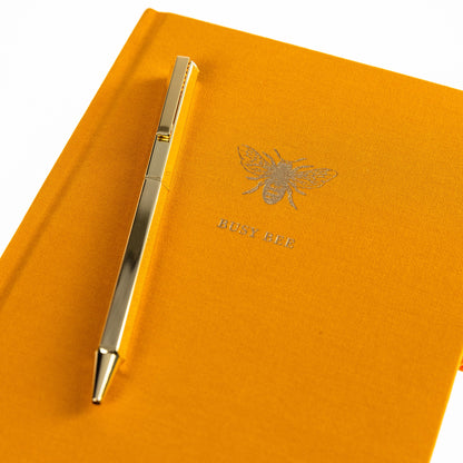 La Petite Presse Bee Fabric-Bound Journal with Pen, A marigold fabric journal with a gold foil bee and the words Busy Bee on the cover, paired with a gold twist pen.
