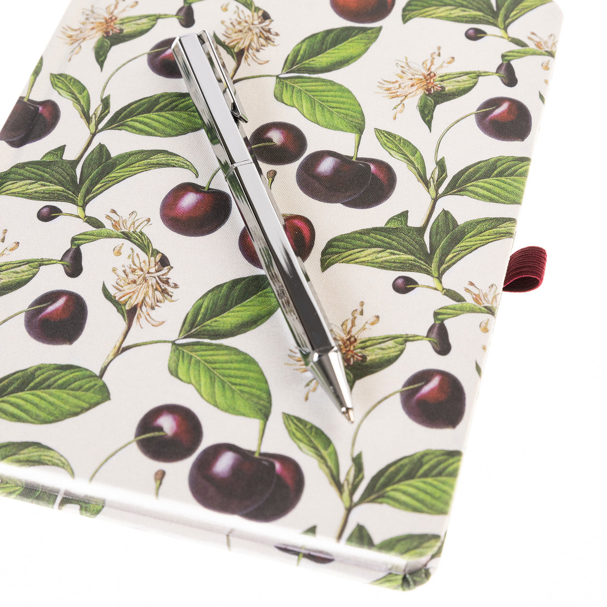 La Petite Presse Cerise Fabric-Bound Journal with Pen, An ivory fabric journal printed with botanical cherries and blossom sprigs, closed with a burgundy elastic band and a silver pen tucked into a loop.