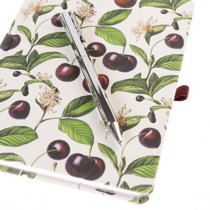 La Petite Presse Cerise Fabric-Bound Journal with Pen, An ivory fabric journal printed with botanical cherries and blossom sprigs, closed with a burgundy elastic band and a silver pen tucked into a loop.