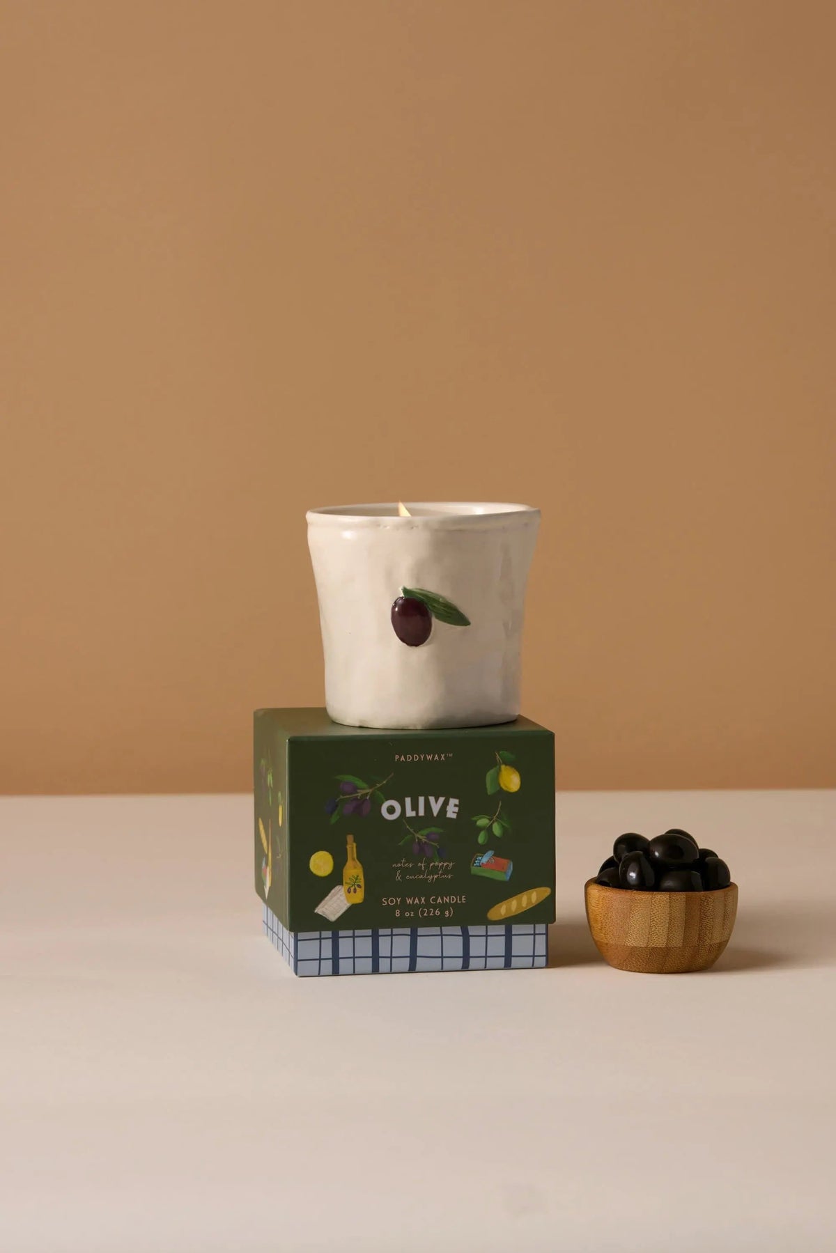 Fig & Olive Bistro Candle 8 oz, A lit ceramic candle with a hand-painted olive and leaf stands on its green Fig and Olive box.