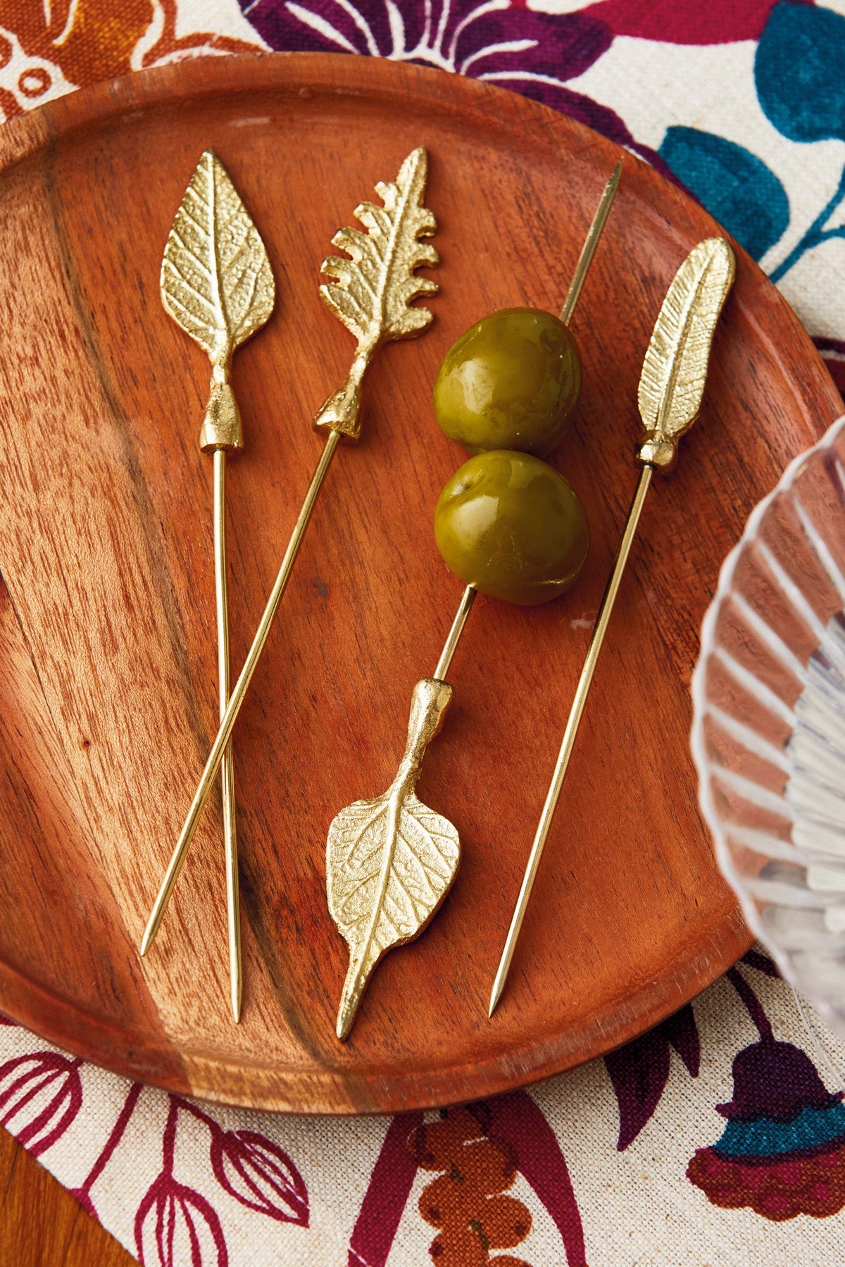 Leaf Cocktail Picks, Set of Four, Four gold metal cocktail picks with different leaf-shaped toppers, including an oak leaf and a feather shape, laid out on a white background.