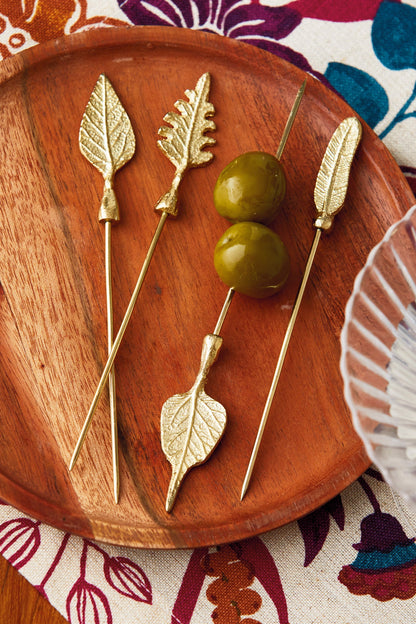 Leaf Cocktail Picks, Set of Four, Four gold metal cocktail picks with different leaf-shaped toppers, including an oak leaf and a feather shape, laid out on a white background.