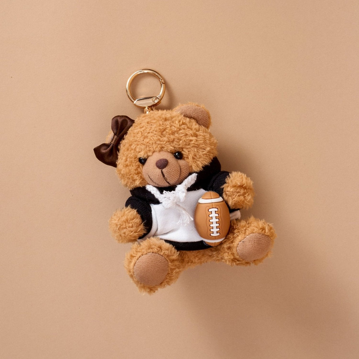 Teddy Bear Bag Charm, A golden brown plush teddy bear bag charm dressed in a black and white football jersey, holding a football, with a gold clasp.