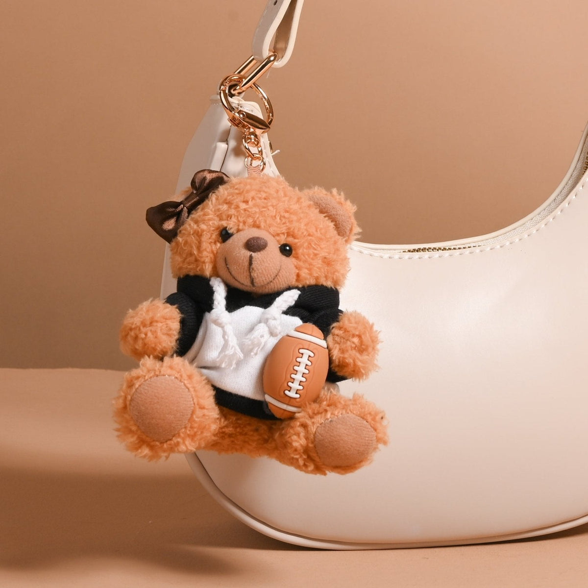 Teddy Bear Bag Charm, A golden brown plush teddy bear bag charm dressed in a black and white football jersey, holding a football, with a gold clasp.