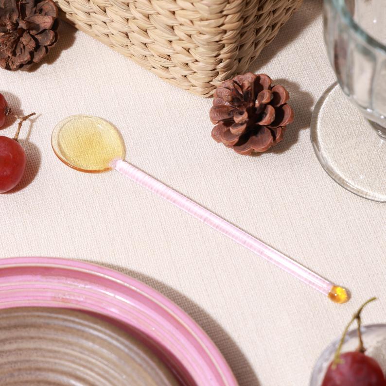 Daisy Glass Stirring Spoon, A glass stirring spoon with a pink handle and a pale yellow bowl, resting on a linen tablecloth near a pink plate and pinecones.