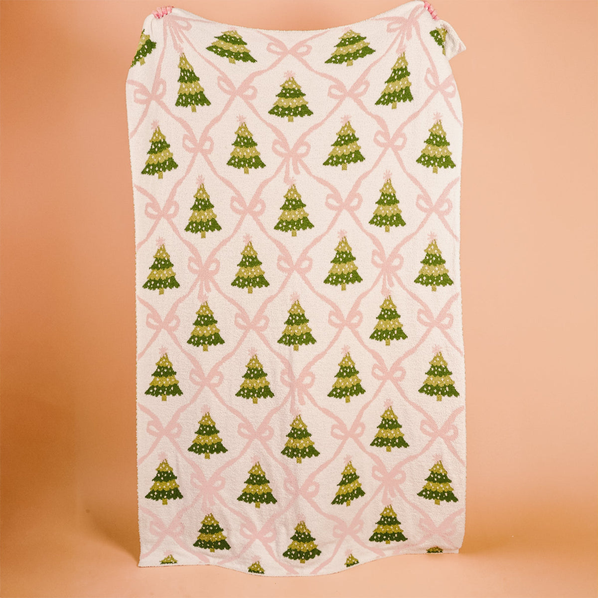 Twinkling Trees Cozy Dreams Luxe Blanket, A cream throw blanket printed with a pattern of green Christmas trees connected by a pink ribbon lattice, held up to show the full design.