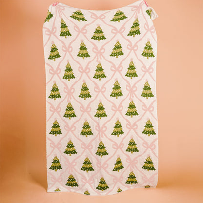 Twinkling Trees Cozy Dreams Luxe Blanket, A cream throw blanket printed with a pattern of green Christmas trees connected by a pink ribbon lattice, held up to show the full design.