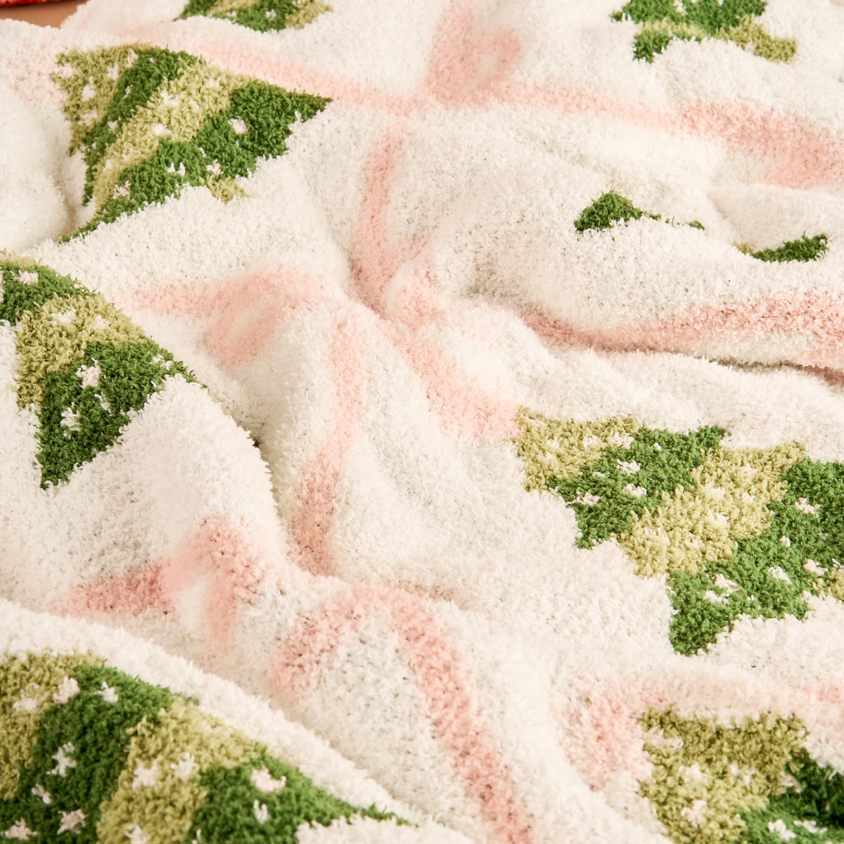 Twinkling Trees Cozy Dreams Luxe Blanket, A cream throw blanket printed with a pattern of green Christmas trees connected by a pink ribbon lattice, held up to show the full design.
