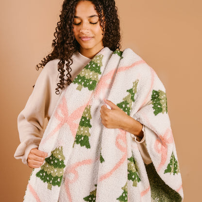 Twinkling Trees Cozy Dreams Luxe Blanket, A cream throw blanket printed with a pattern of green Christmas trees connected by a pink ribbon lattice, held up to show the full design.
