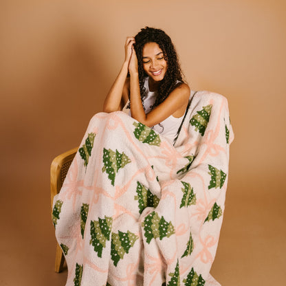 Twinkling Trees Cozy Dreams Luxe Blanket, A cream throw blanket printed with a pattern of green Christmas trees connected by a pink ribbon lattice, held up to show the full design.