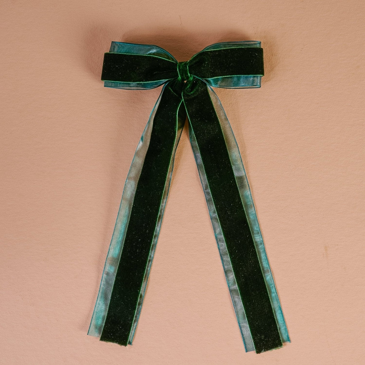 Velvet Luxe Layered Hair Bow, A deep green velvet bow with long tails and a sheer edge, laid flat against a pink background.