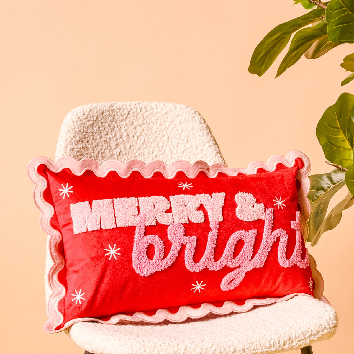 Scalloped Rectangle Throw Pillow, A blush pink lumbar pillow with three tufted green Christmas trees tied in red bows and a pink scalloped edge, set on a boucle chair.