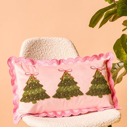 Scalloped Rectangle Throw Pillow, A blush pink lumbar pillow with three tufted green Christmas trees tied in red bows and a pink scalloped edge, set on a boucle chair.