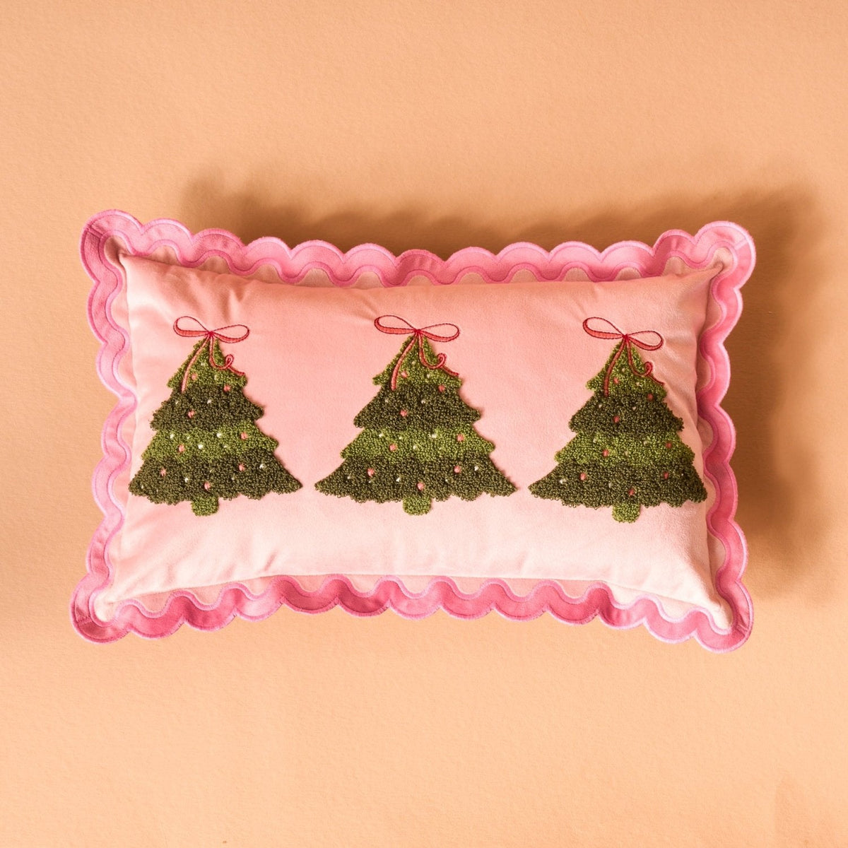 Scalloped Rectangle Throw Pillow, A blush pink lumbar pillow with three tufted green Christmas trees tied in red bows and a pink scalloped edge, set on a boucle chair.