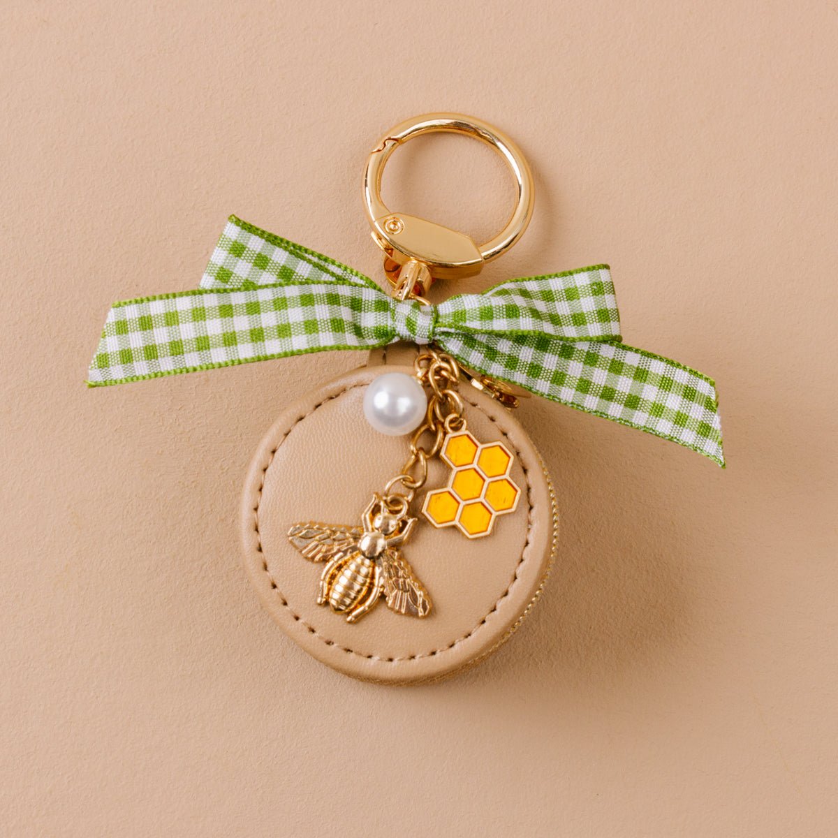 Travel Jewelry Case Bag Charm, A tan round zip case charm with a green gingham bow, a pearl, a gold bee, and a honeycomb charm, in the Hey Honey design.